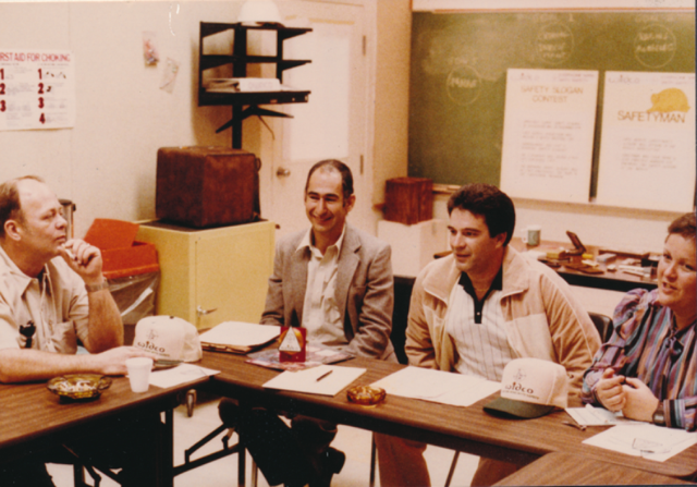 Four adults sit around a classroom table covered with papers and coffee cups, with a chalkboard and posted notices behind them.